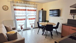 Ein Wohnzimmer mit einer Couch, einem Esstisch mit Stühlen, einem an der Wand montierten Fernseher und einem Kühlschrank.