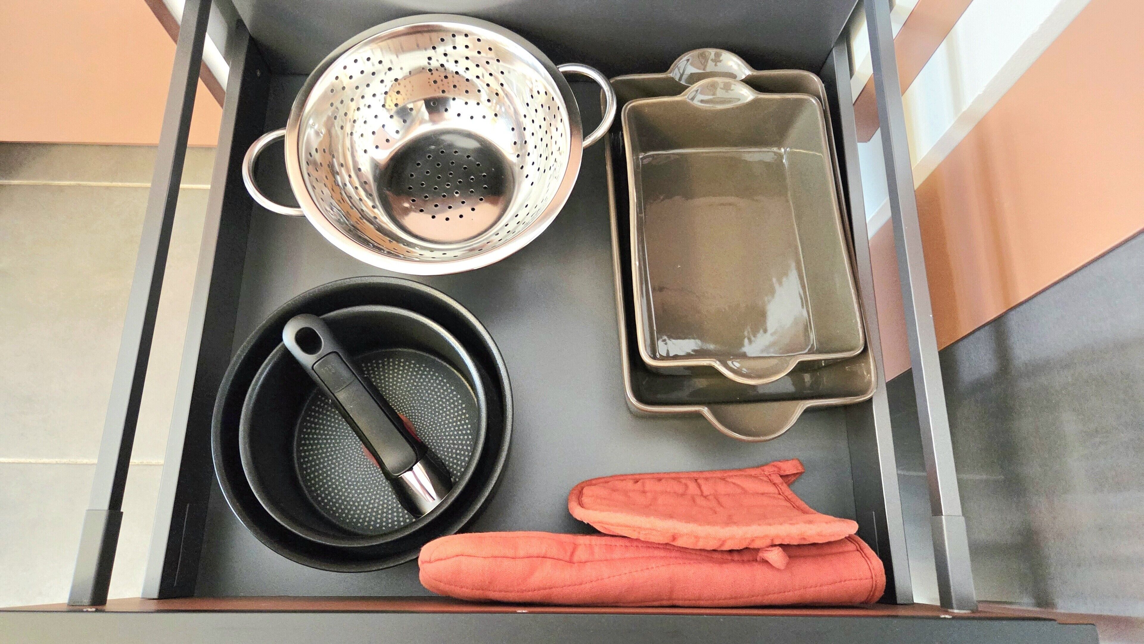 A kitchen drawer with a colander, a pan, a pot, and oven mitts.