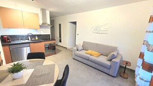 Eine moderne Küche mit Geschirrspüler, Einbauofen und Herd. Ein graues Sofa mit gelbem Kissen und ein Schild mit der Aufschrift 'Welcome' sind an der Wand zu sehen. Ein Esstisch mit Stühlen ist ebenfalls sichtbar.