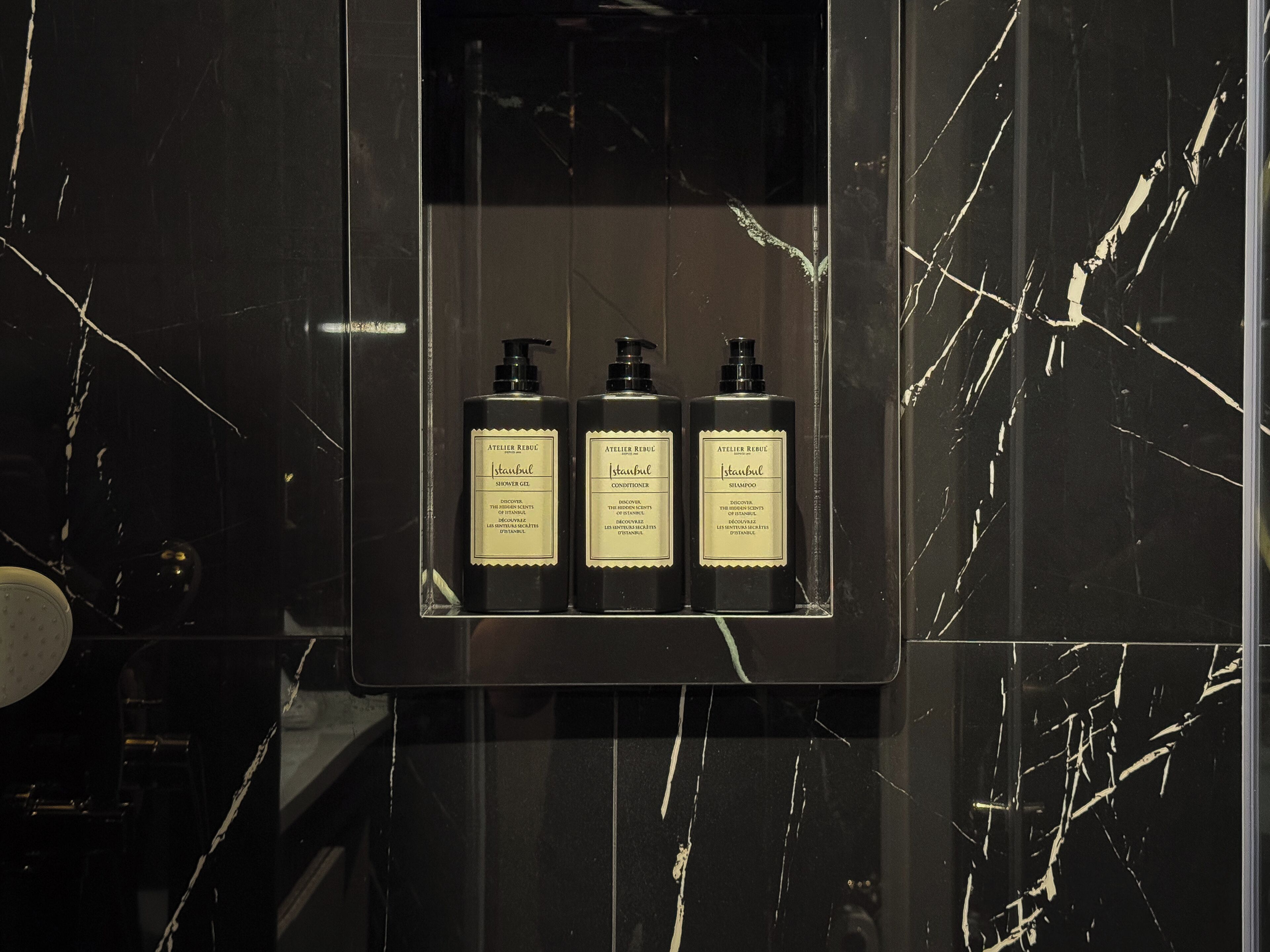 Ein Badezimmer mit schwarzem Marmor an der Wand und einem Regal mit drei Flaschen Duschgel, Conditioner und Shampoo.