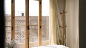 A bedroom with a bed, a hanging light fixture, a window with curtains, and a balcony with a metal railing. - The Lodge (Aourir)