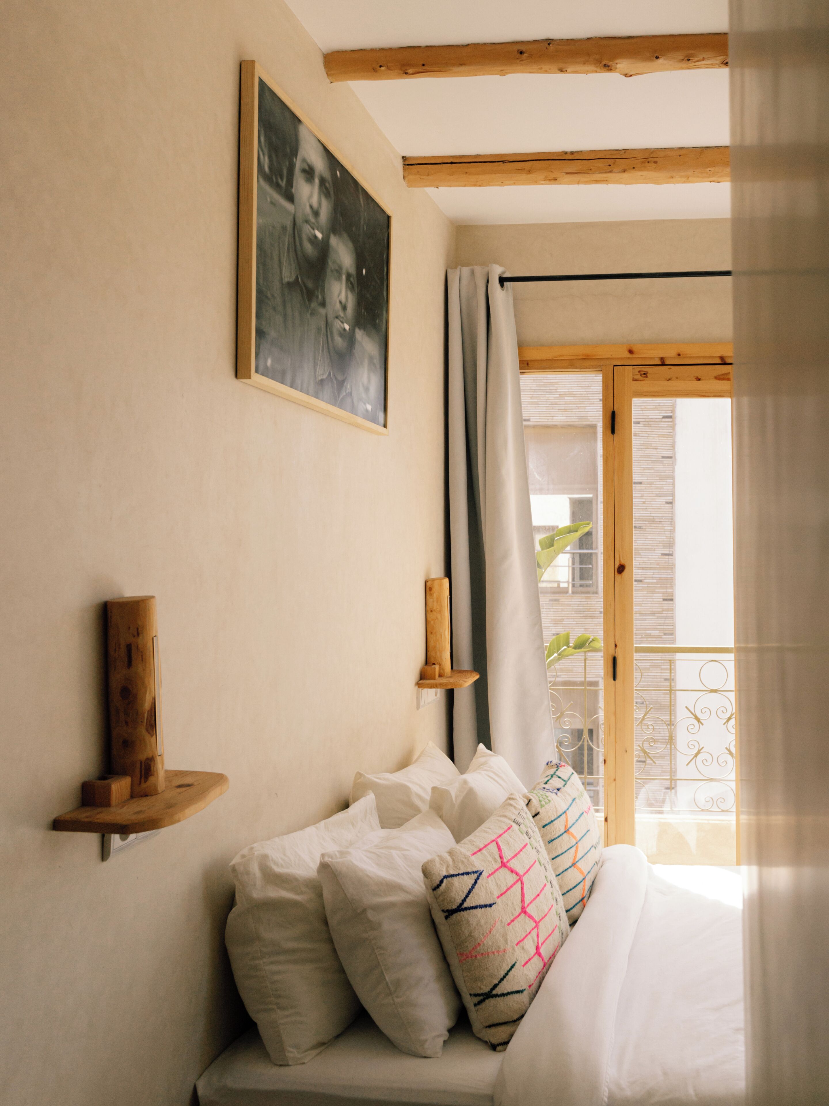 A bedroom with a bed, pillows, a wooden shelf, a framed picture, and a window with curtains.