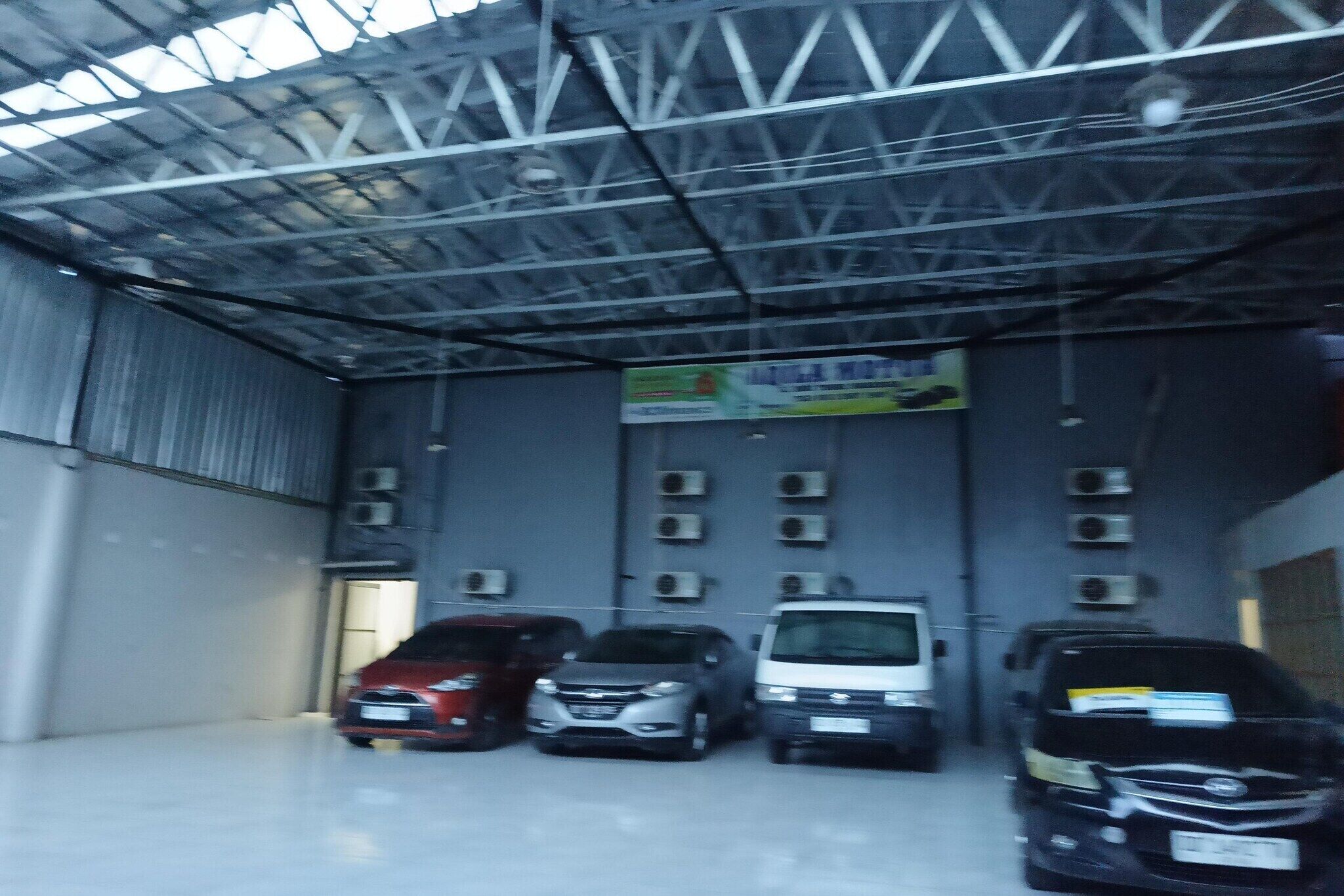 Un espace de stationnement couvert où sont garées plusieurs voitures, avec des unités de climatisation fixées aux murs et un panneau suspendu au plafond.