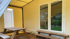 A wooden deck with a picnic table and benches, a sliding glass door, and a white curtain. - Cozy Coco Sweet Glamping Tent for 4 (Rijkevorsel)