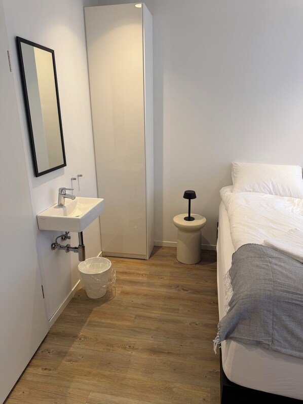 A bedroom with a bed, a bedside table with a lamp, a white sink, and a mirror.