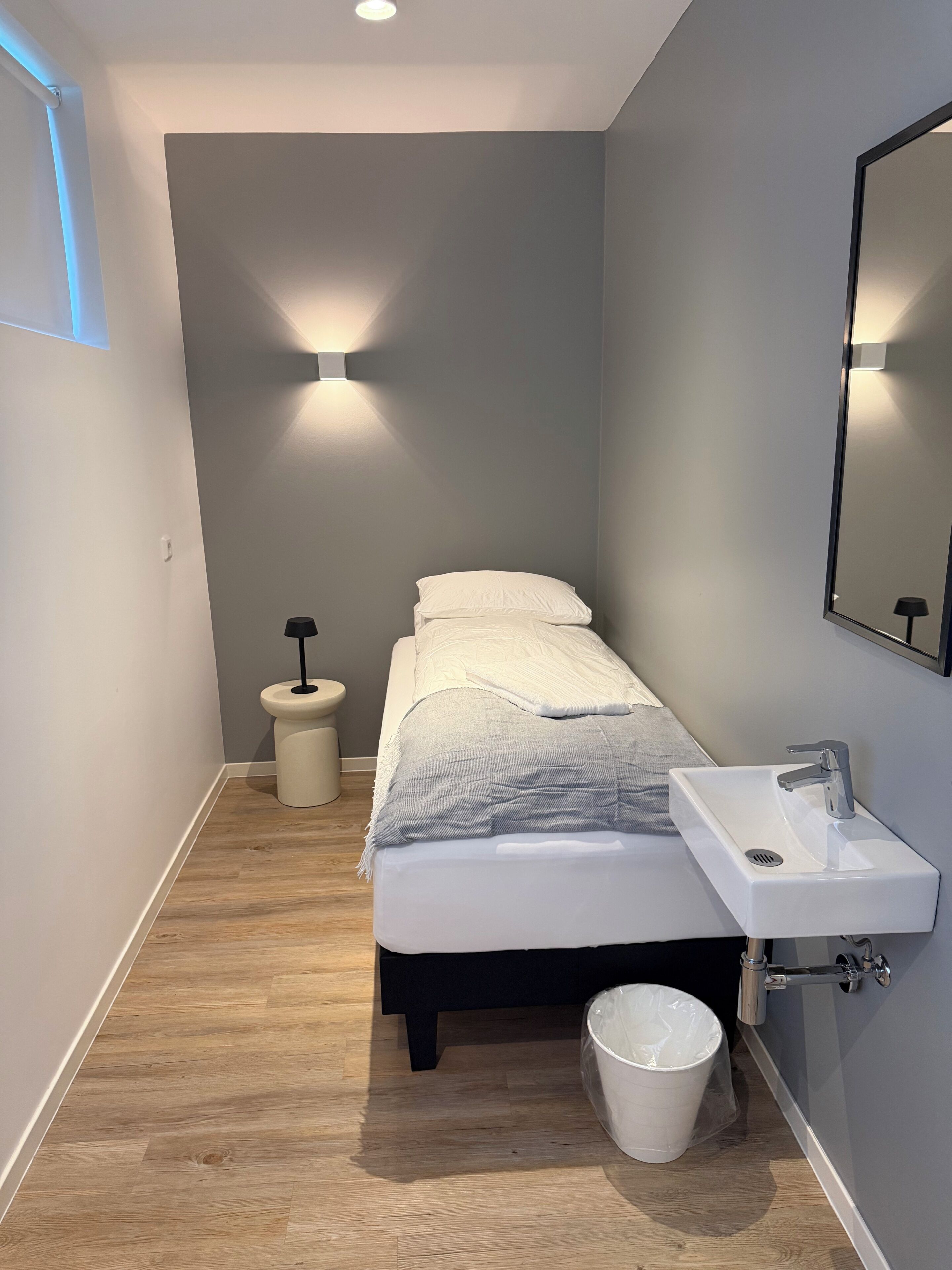A narrow hotel room with a bed, a bedside table, a small stool, a white bucket, and a wall-mounted sink.