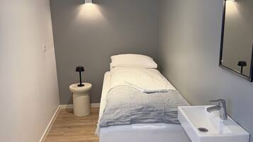 A narrow hotel room with a bed, a bedside table, a small stool, a white bucket, and a wall-mounted sink.