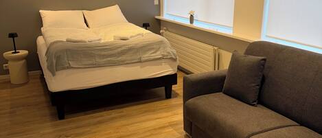 Une chambre à coucher avec un lit, un canapé gris, un plancher en bois et des lampes fixées au mur.