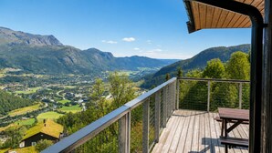 Terraza de madera con vista a un valle, montañas y un edificio pequeño.