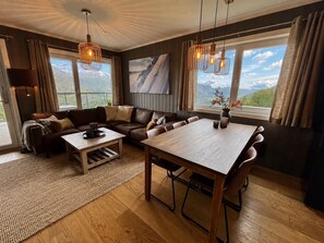 A spacious living area with a wooden dining table, leather sofas, and a large window offering a mountain view. - Mountain gem-  Ski in/out & panorama views (Hemsedal)