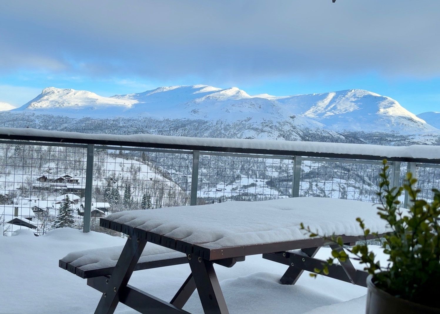 Un espacio exterior cubierto de nieve con una mesa y sillas de metal, una valla metálica y una vista de montañas nevadas al fondo.
