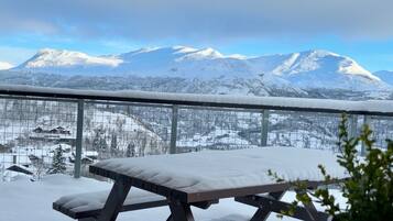 Un espacio exterior cubierto de nieve con una mesa y sillas de metal, una valla metálica y una vista de montañas nevadas al fondo.