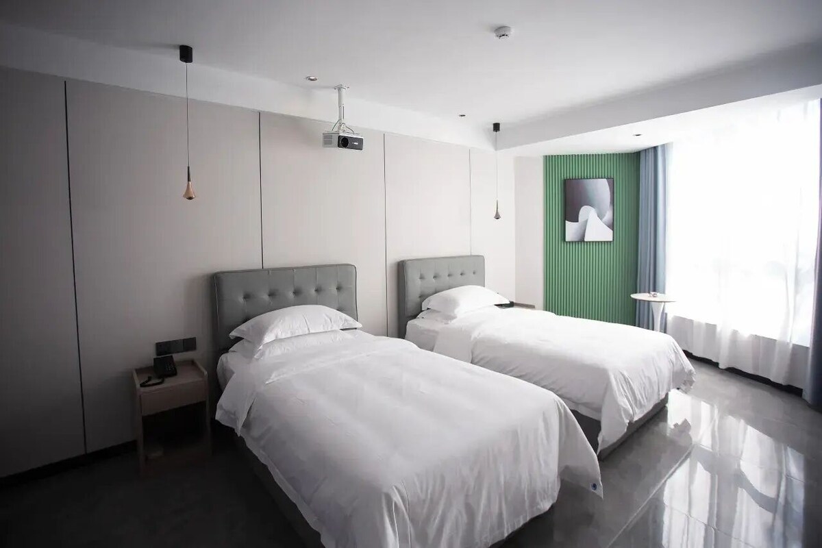 A modern hotel room with two beds, a small bedside table, a projector, and a green accent wall.