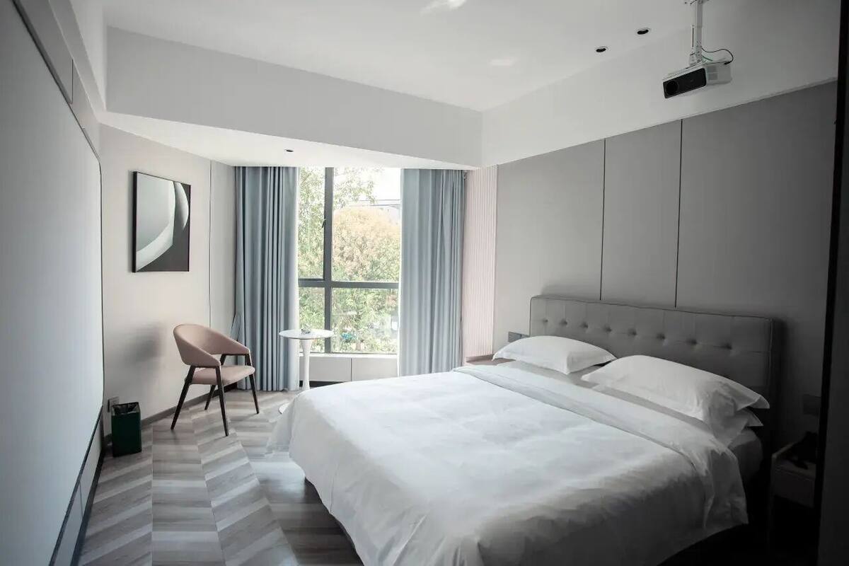 A modern hotel room with a large bed, a chair, a small table, and a window with curtains.