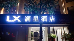 Entrada de hotel con un letrero iluminado llamativo, puerta de vidrio y plantas en macetas afuera.
