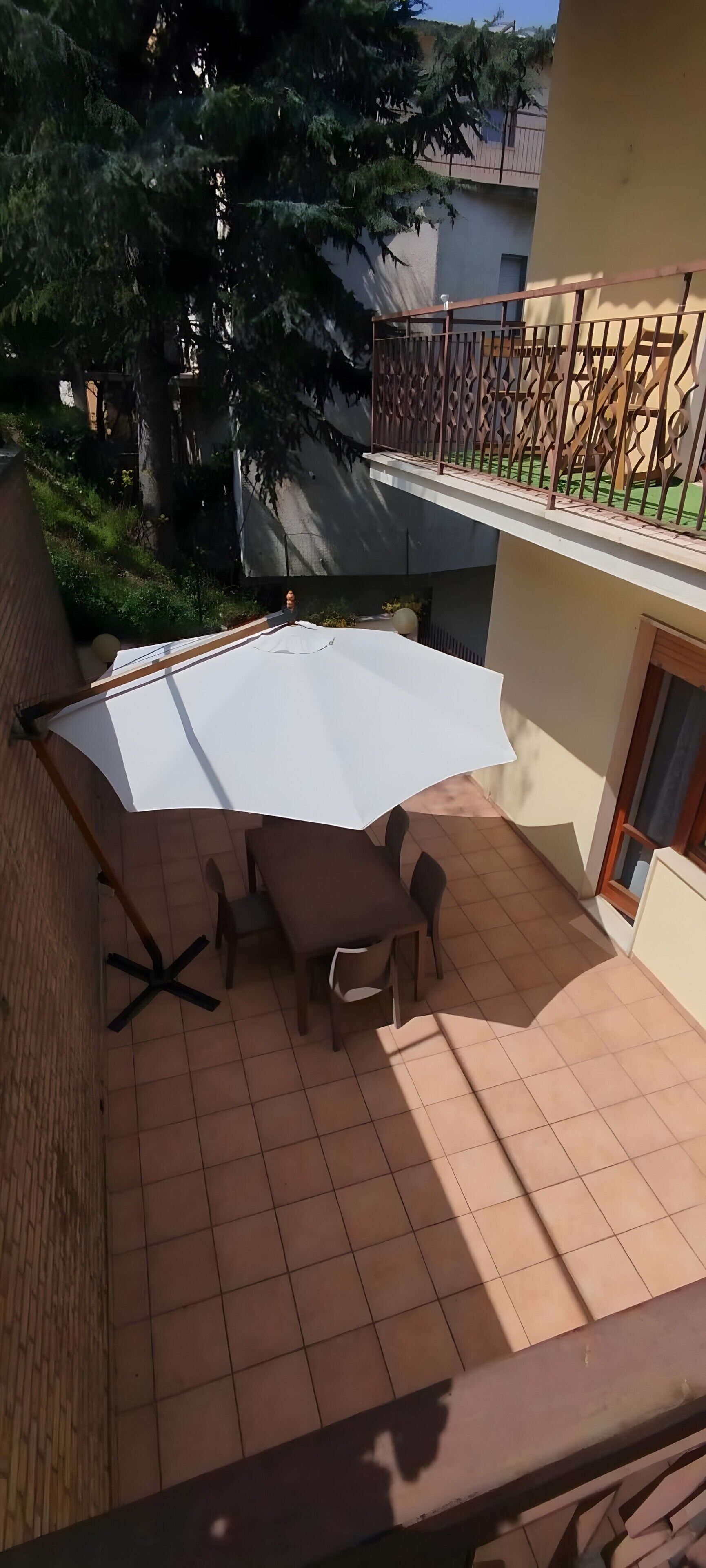 Une terrasse avec une table et des chaises, un parasol blanc, et une vue sur des arbres et un bâtiment.