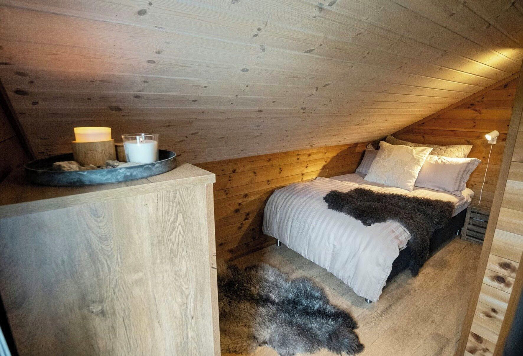 Um quarto de sótão aconchegante com teto de madeira, cama com roupa de cama branca, tapete de pele e uma pequena mesa com velas.