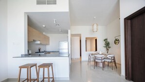 A modern kitchen with a central island, wooden stools, a dining area with a table and chairs, and a dark wooden door. - Bloomfields 1BR in Marina Bay 1  (Abu Dhabi)