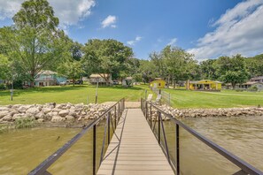 Eine Holzbrücke führt zu einer Wiese mit Häusern und einem Teich.