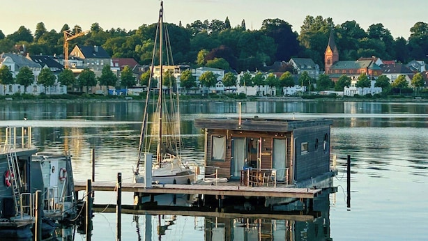 Une péniche flottante est amarrée sur une étendue d’eau calme, sous un ciel dégagé, et on aperçoit au loin le clocher d’une église.