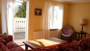 Ein Zimmer mit Holztisch, einem stuhlförmigen Sessel mit Blumenmuster, einer Couch und Blick durch ein Fenster mit weißem Vorhang.