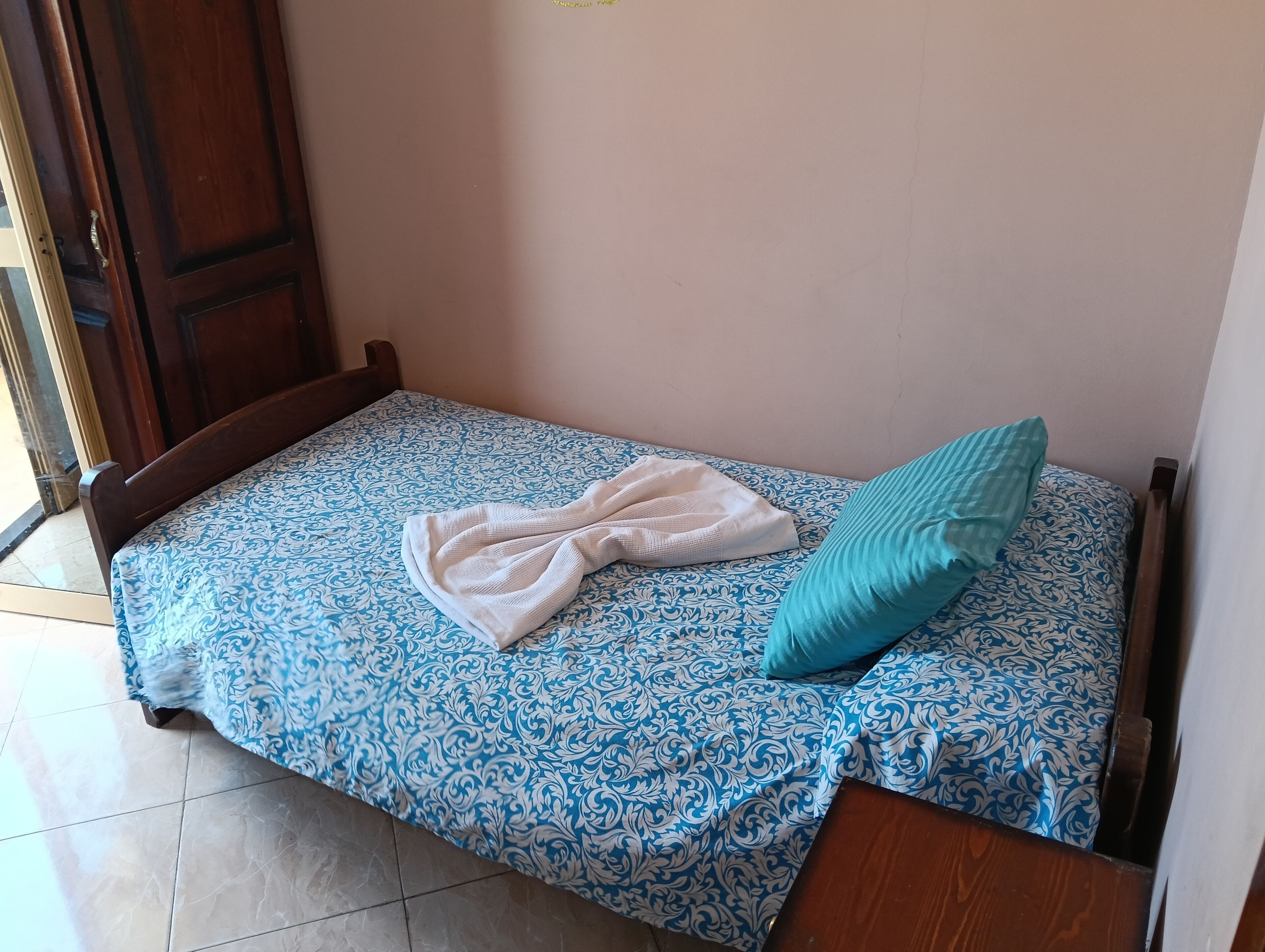Una cama pequeña con una colcha estampada, una toalla blanca y un cojín azul verdoso.