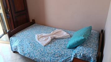Una cama pequeña con una colcha estampada, una toalla blanca y un cojín azul verdoso.