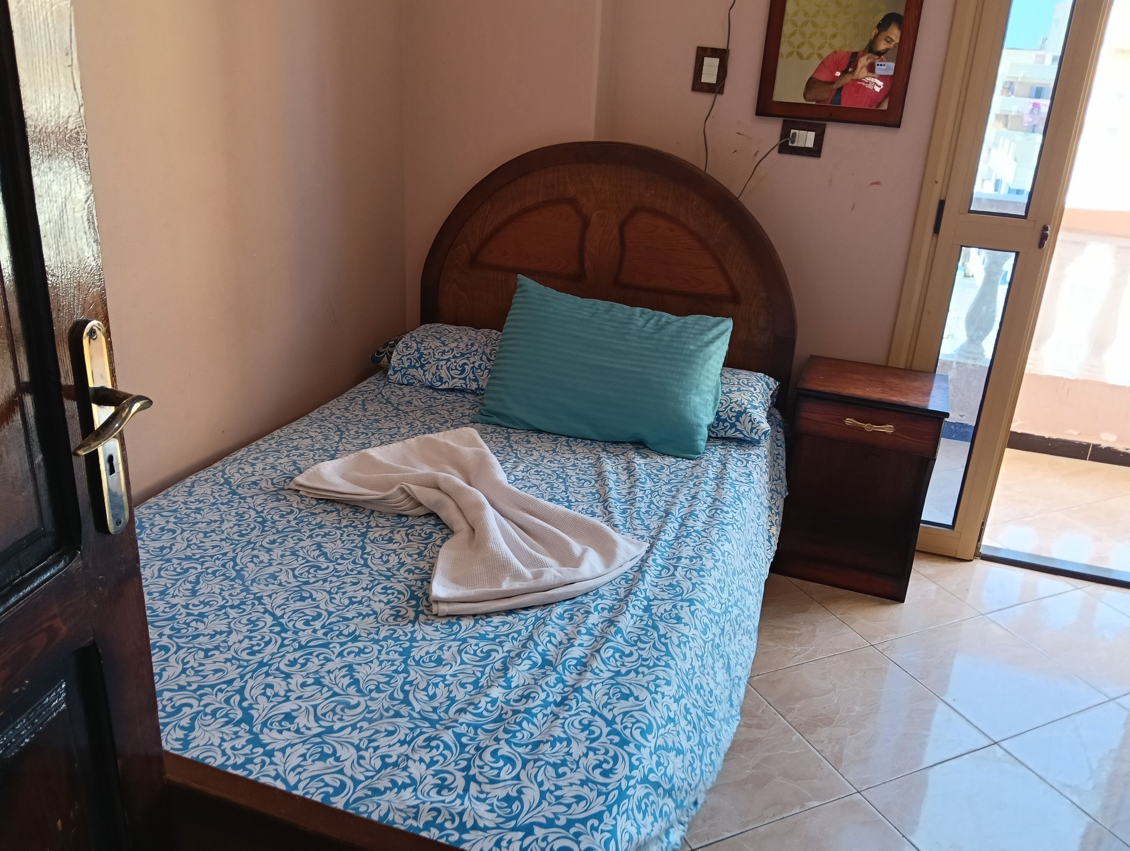 Ein ordentlich bezogenes Bett mit gemusterten Betttextilien, ein Holzkopfende, ein Nachttisch und ein Spiegel an der Wand.