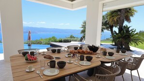 Ett matbord dukat med frukostmat, omgivet av stolar, med utsikt över havet och bergen.