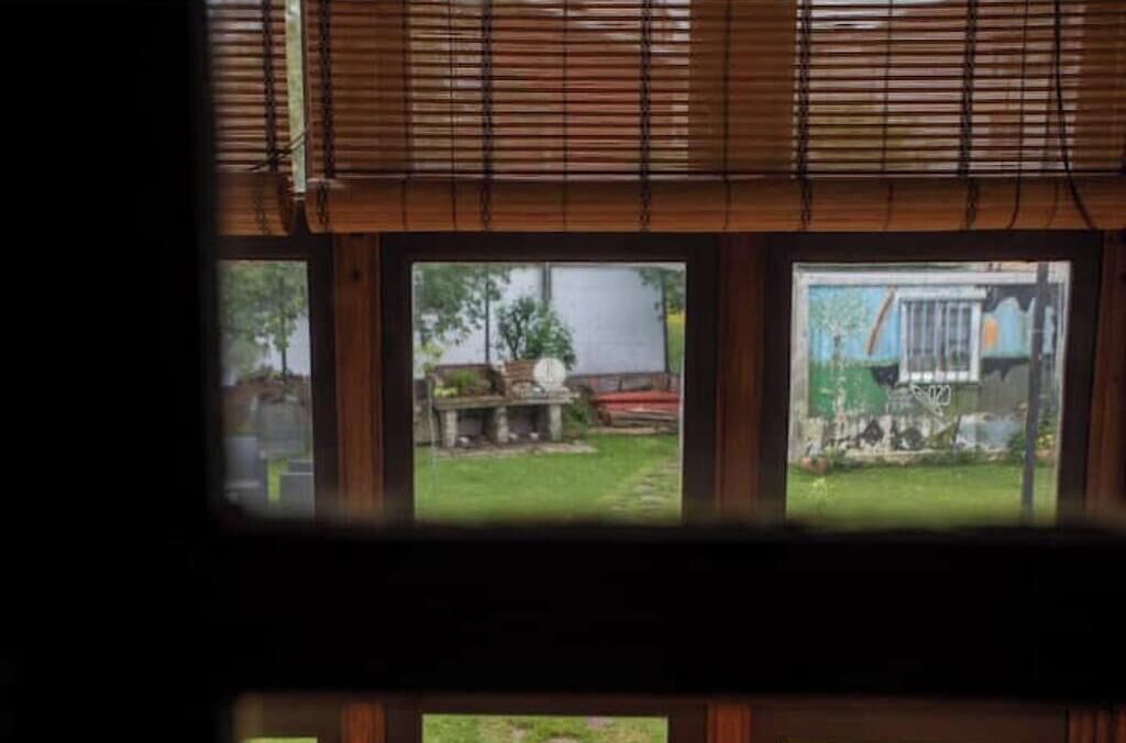 A room with three windows, wooden blinds, and a view of a yard with a fence and some greenery.