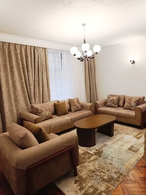 A living room with a sofa set, a coffee table, a chandelier, and a patterned rug. - meltonia suites by extended stays (Nairobi)