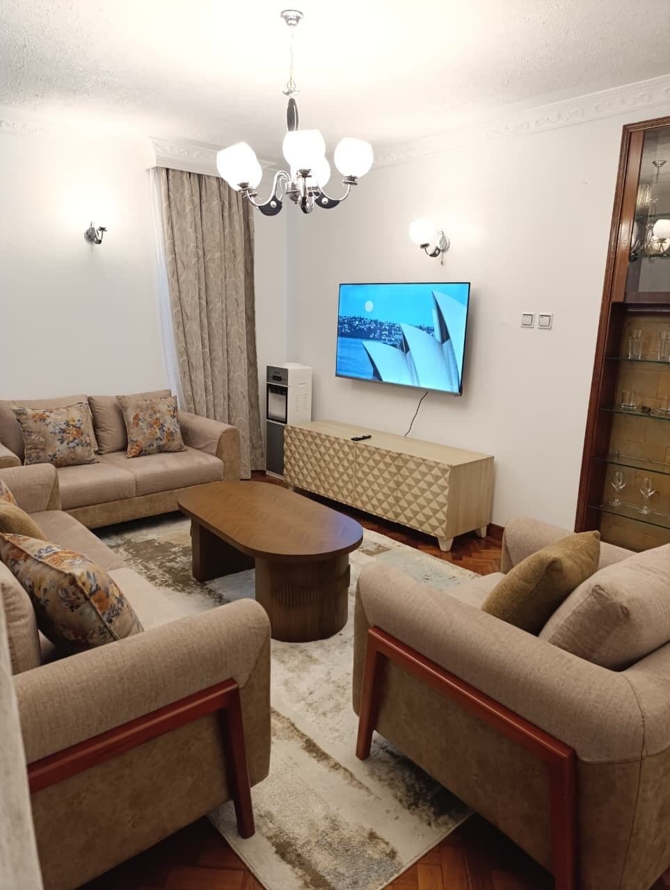 A living room with a sofa set, a coffee table, a TV mounted on the wall, a chandelier, and a cabinet with glass doors.