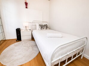 A bedroom with a white bed, a bedside table with a lamp, a small decorative vase, and a round rug on a wooden floor. - 3BDR Apt in the Heart of Strathfield (Strathfield)