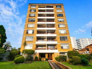 A multi-story residential building with a central entrance, surrounded by well-maintained greenery and a clear sky. - 3BDR Apt in the Heart of Strathfield (Strathfield)