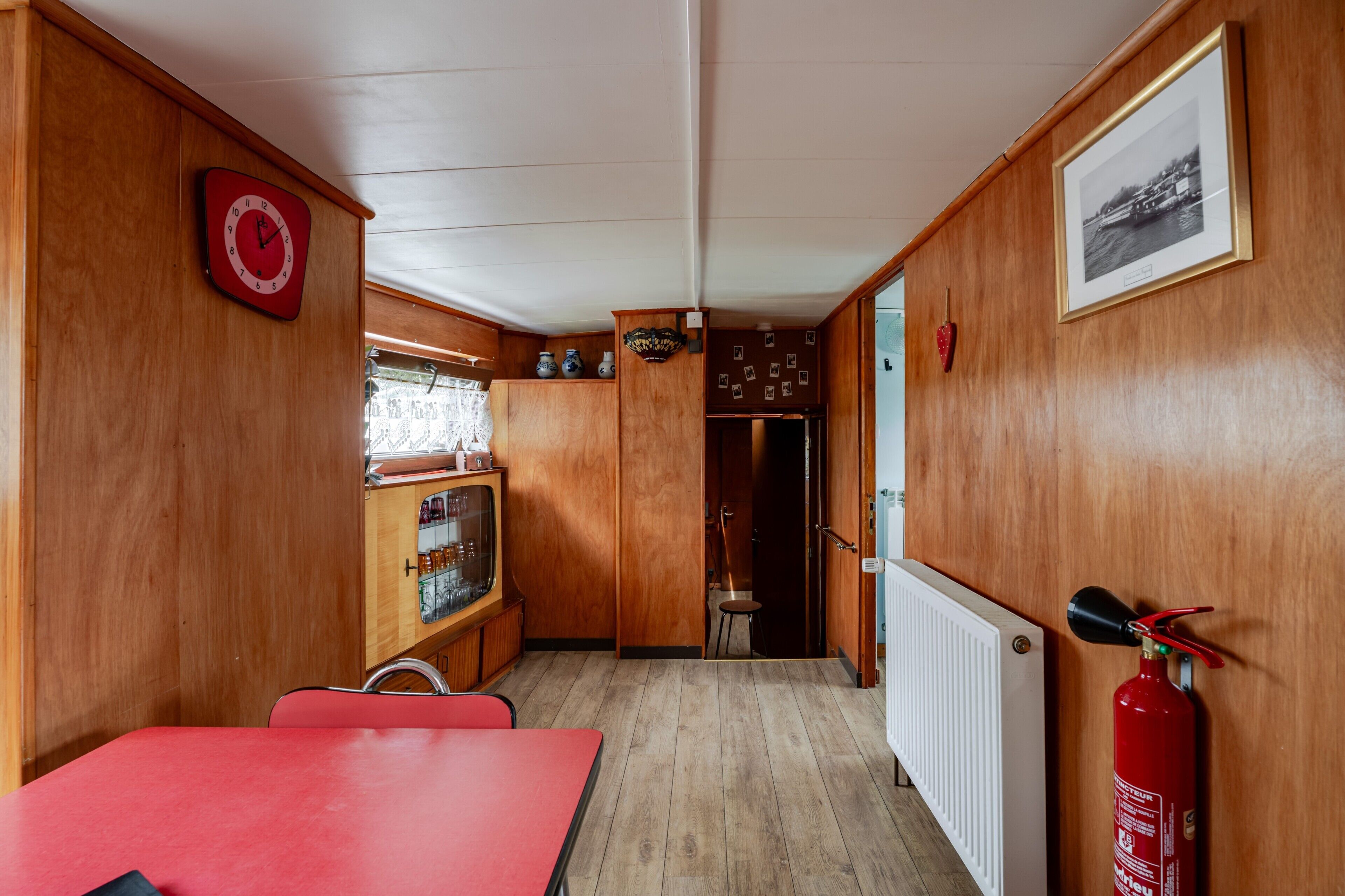 A wooden interior with a red table, a red clock, and a framed photograph on the wall.