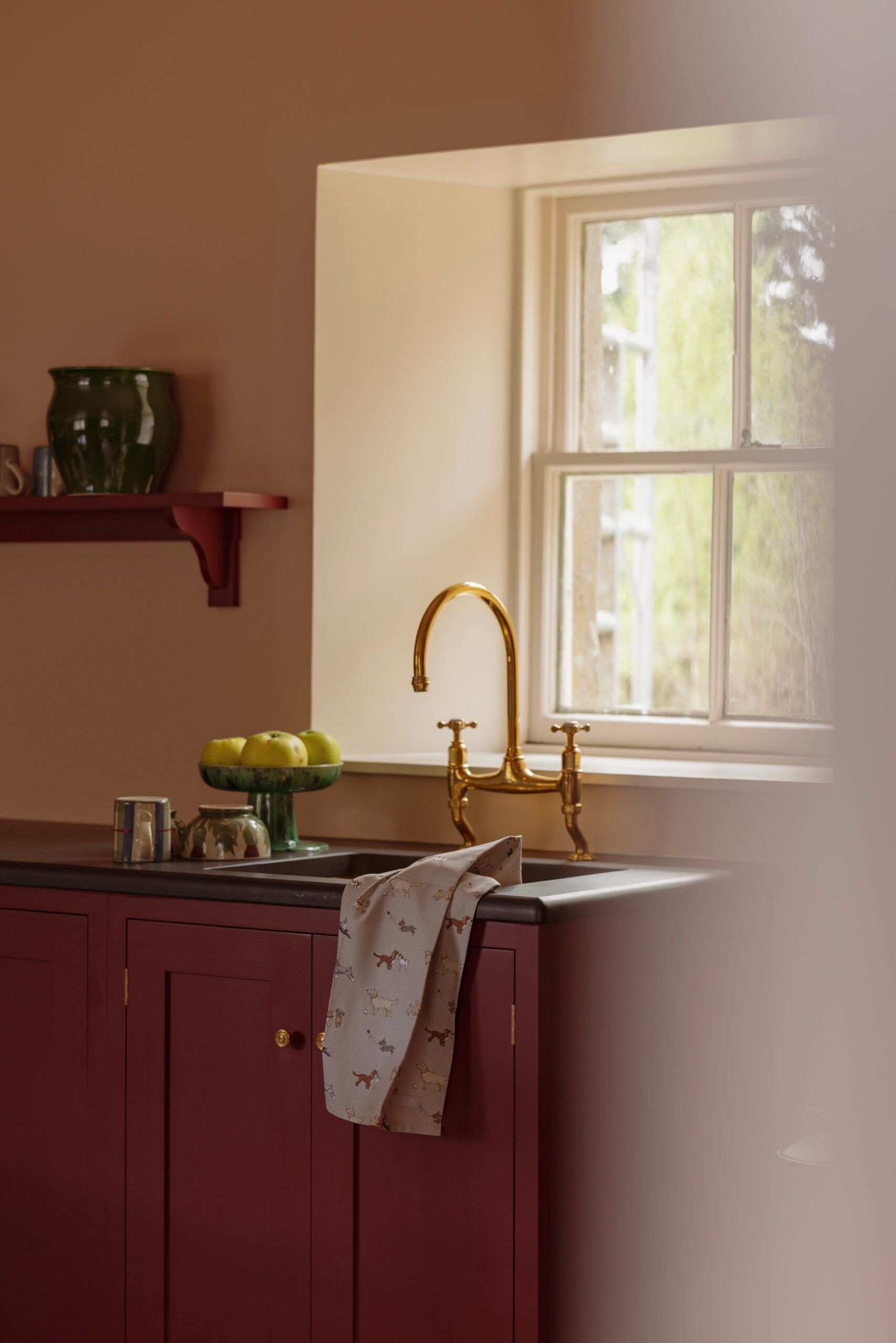 A kitchen with a window, a sink with a gold faucet, a bowl of apples, and a red cabinet.