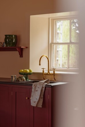 A kitchen with a window, a sink with a gold faucet, a bowl of apples, and a red cabinet. - Moy Cottage - sleeps 10, 5 bedrooms, pet-friendly Highland retreat (Moy)