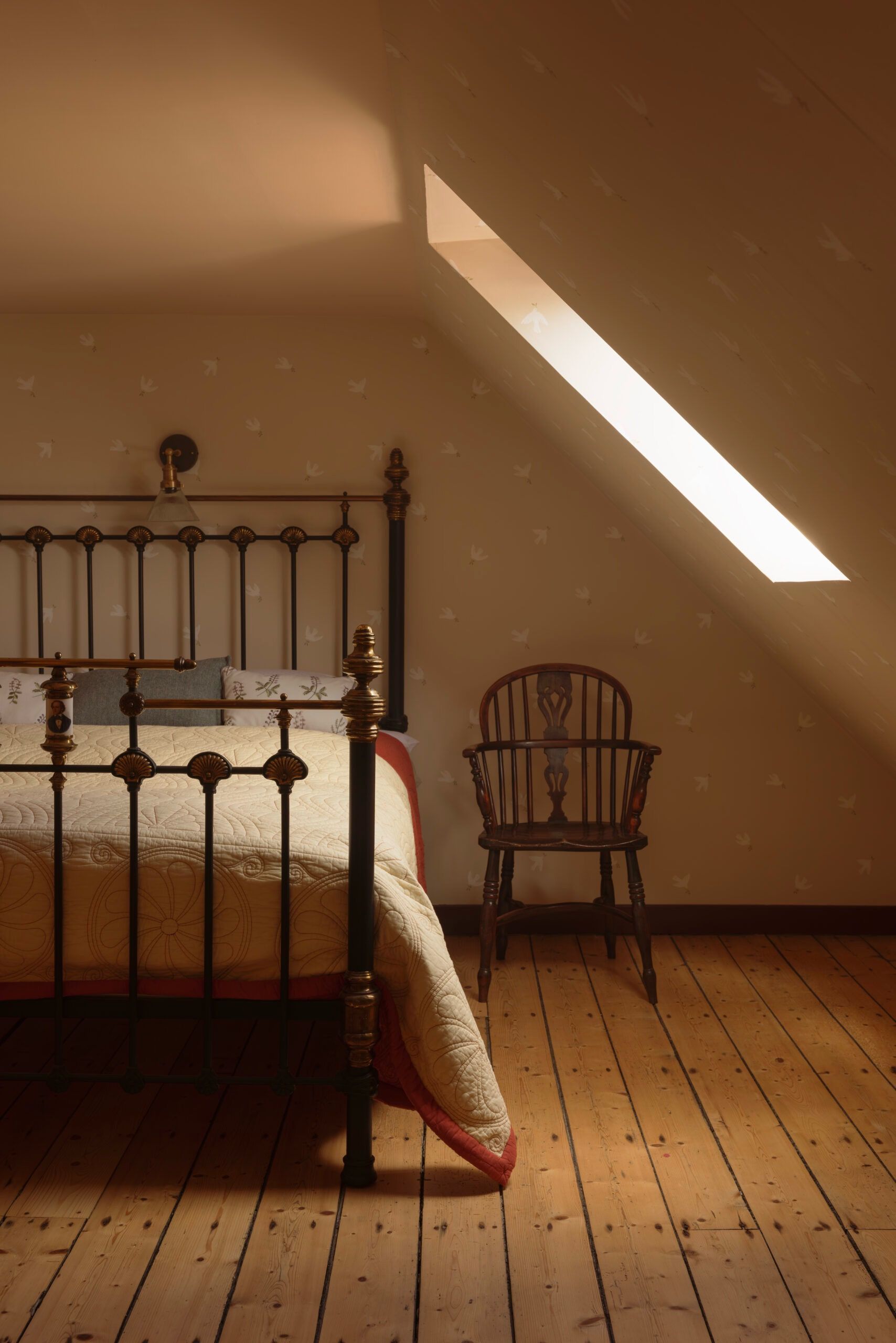 A bedroom with a sloped ceiling, a bed with a headboard, a wooden chair, and a skylight.