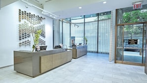 Uma área de recepção moderna, com entrada por porta de vidro, painel de cores fixado na parede e balcão de atendimento com computador e planta.