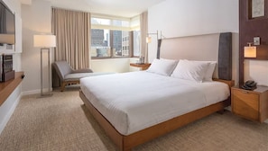 Quarto de hotel com uma cama grande, uma poltrona, uma escrivaninha, um abajur e uma televisão.