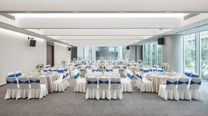 Un amplio salón de conferencias con varias mesas redondas, cintas blancas y azules en las sillas y grandes ventanales con vistas al exterior.