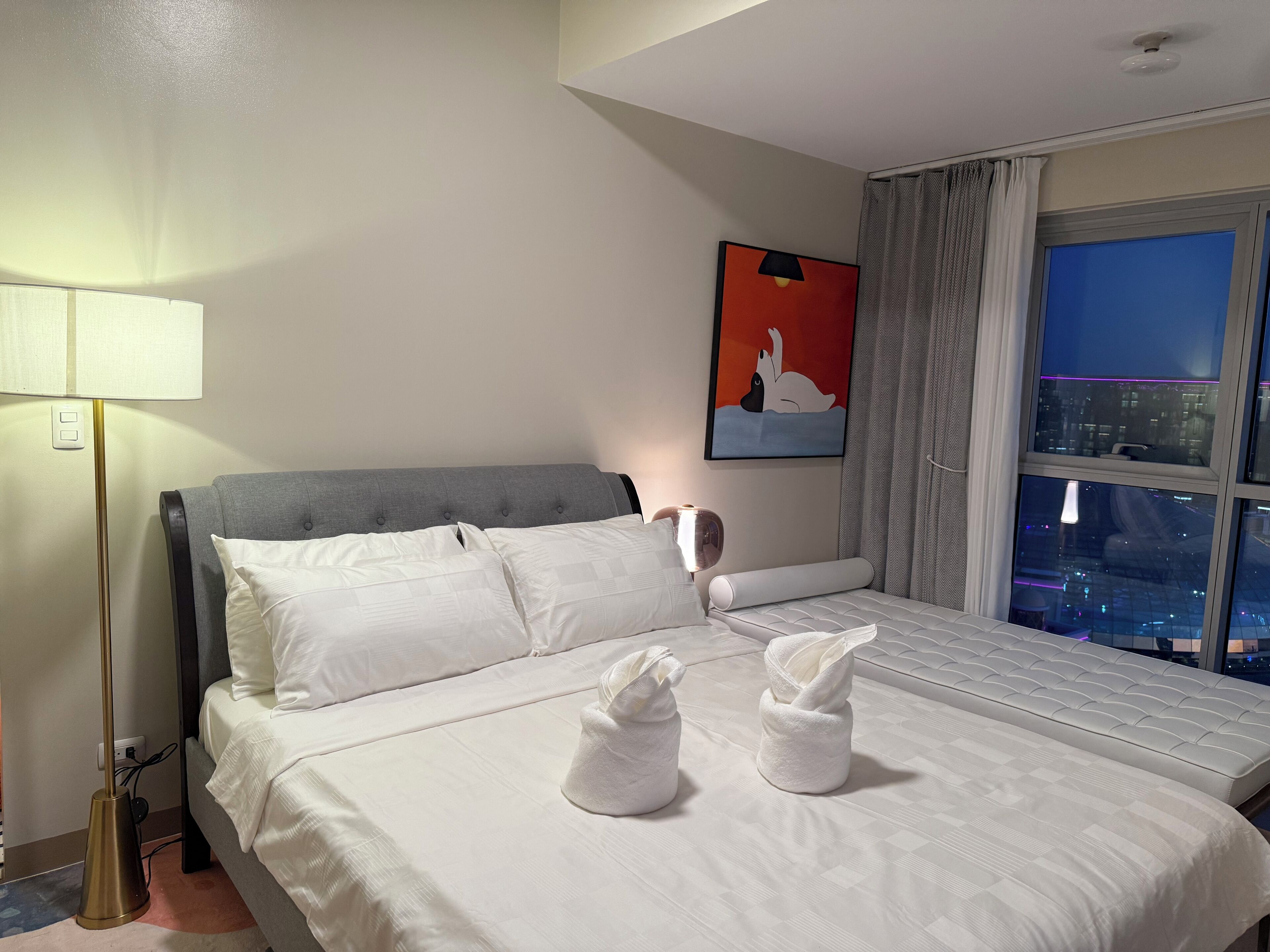 A hotel room with a bed, a lamp, a painting, and a window with a city view.