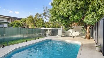 Ein rechteckiger Pool mit Holzterrasse, umgeben von einem Metallzaun und einem Baum, der Schatten spendet.