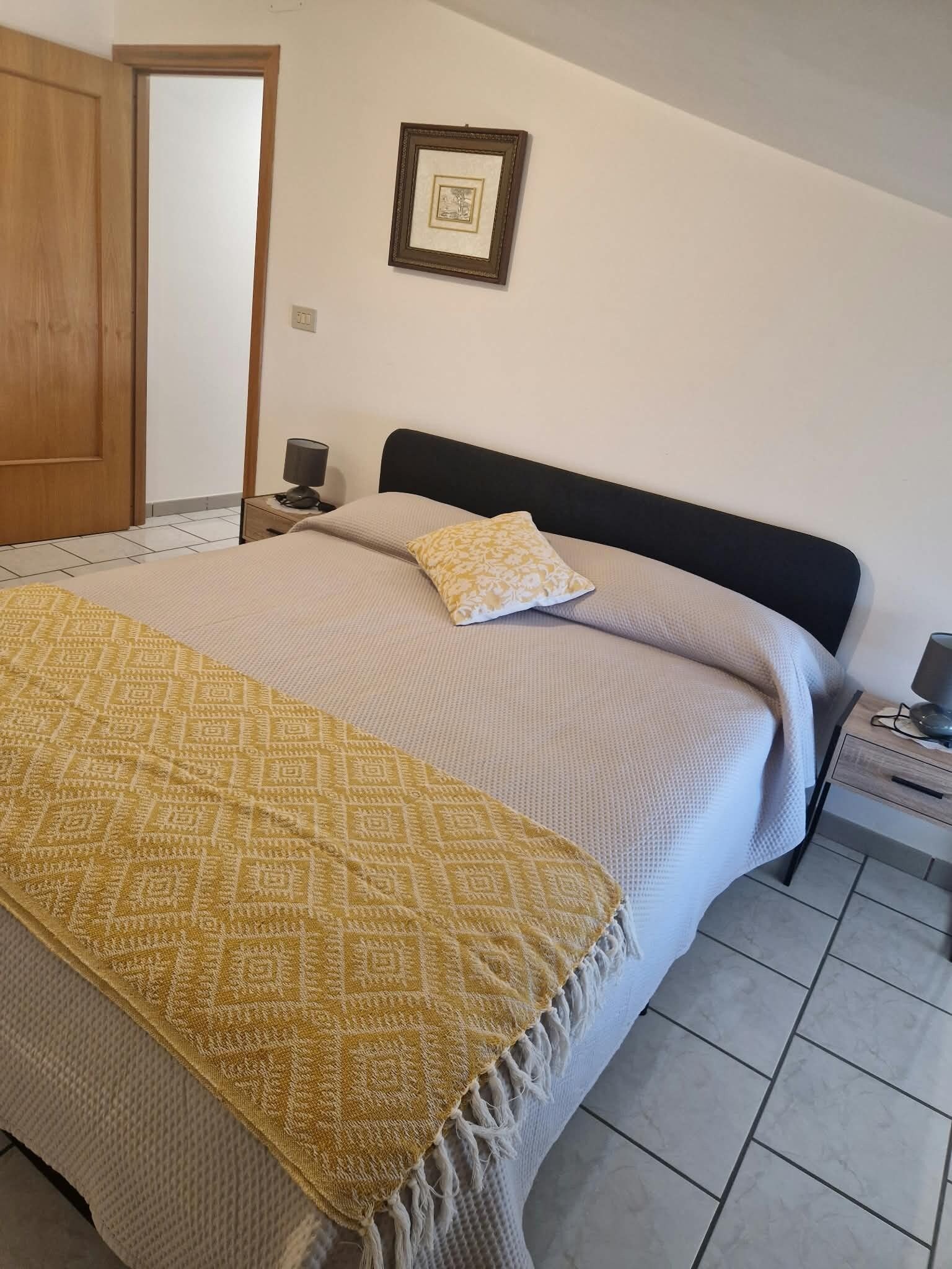 A bedroom with a bed, a bedside table, a lamp, a picture on the wall, and a door.