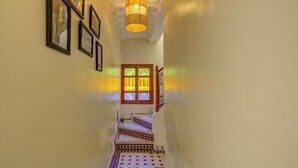 Um corredor com ladrilhos estampados, uma escada com grades decorativas e um candeeiro fixado na parede.