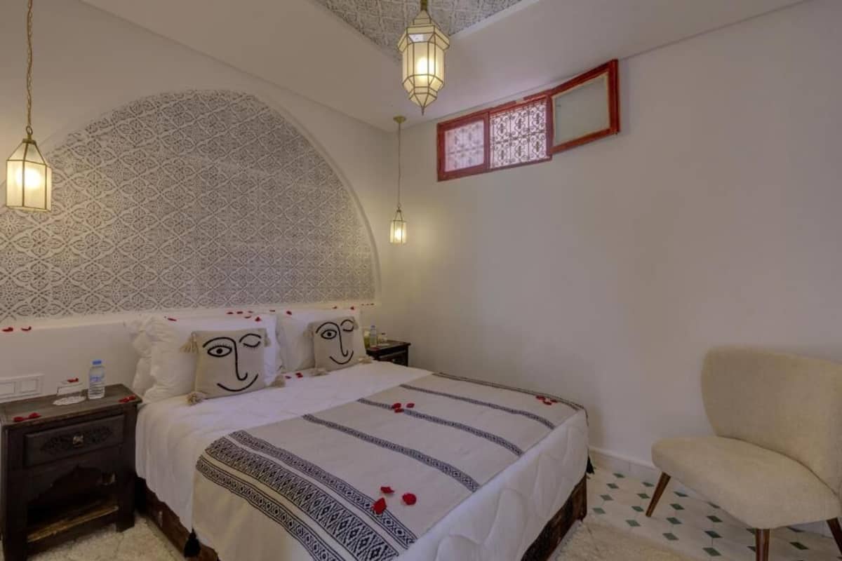 A bedroom with a patterned wall, a bed with a patterned bedspread, a nightstand, a chair, and a decorative mirror.