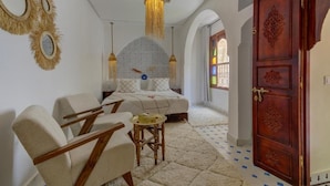 Quarto com cama, duas cadeiras, uma mesinha, espelho decorativo, luminária pendente e guarda-roupa de madeira.