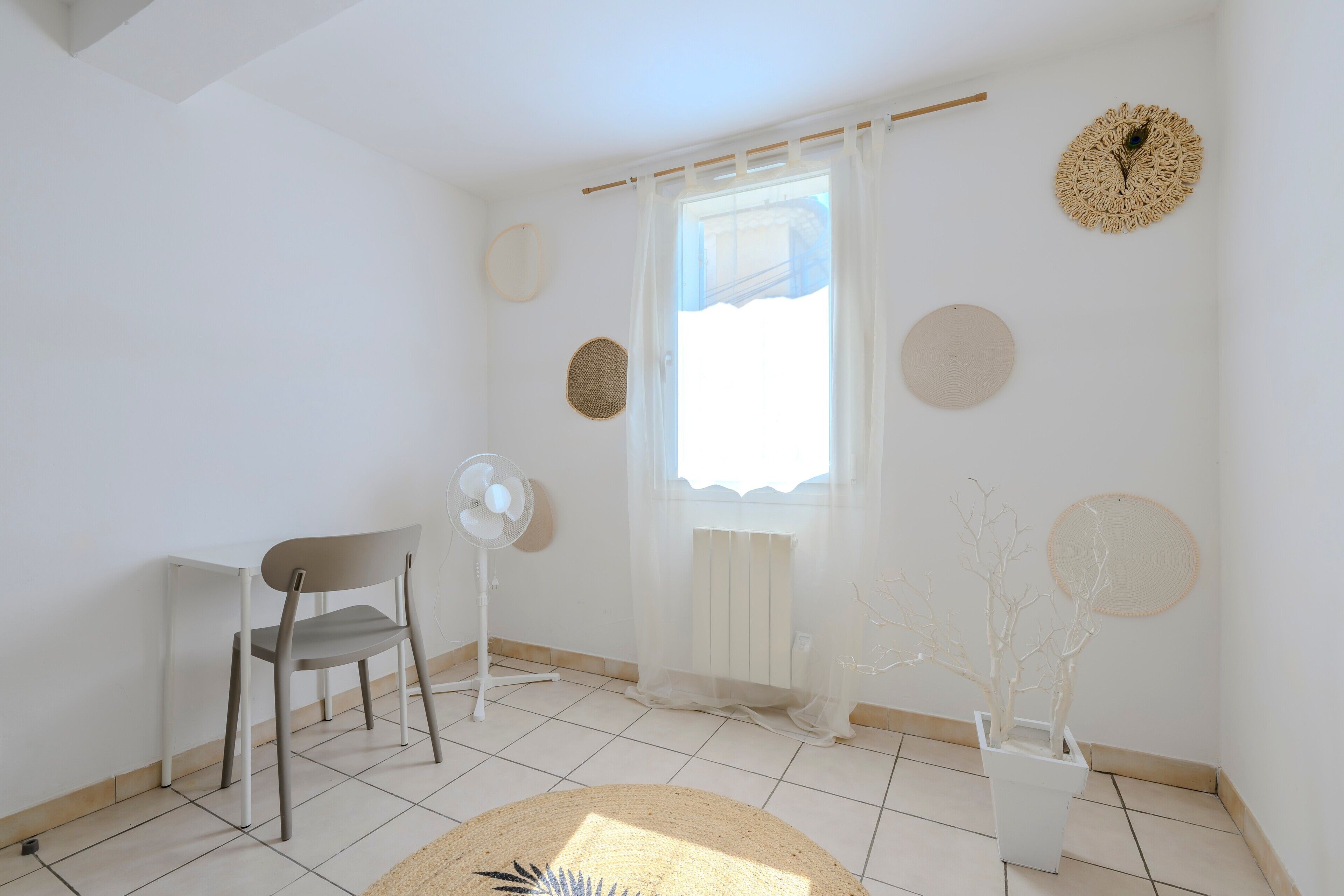 Une pièce comprenant une chaise, un ventilateur, une fenêtre avec des rideaux légers et des éléments décoratifs sur les murs.