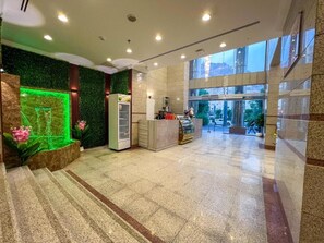 Vestíbulo de hotel con fuente iluminada en verde, una recepción y vistas al exterior a través de amplios ventanales.