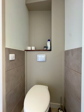 A small bathroom with a toilet, a shelf with toiletries, and a light switch. - L'Ajax, 2-bedroom apartment in the city center and by the sea (Ajaccio)